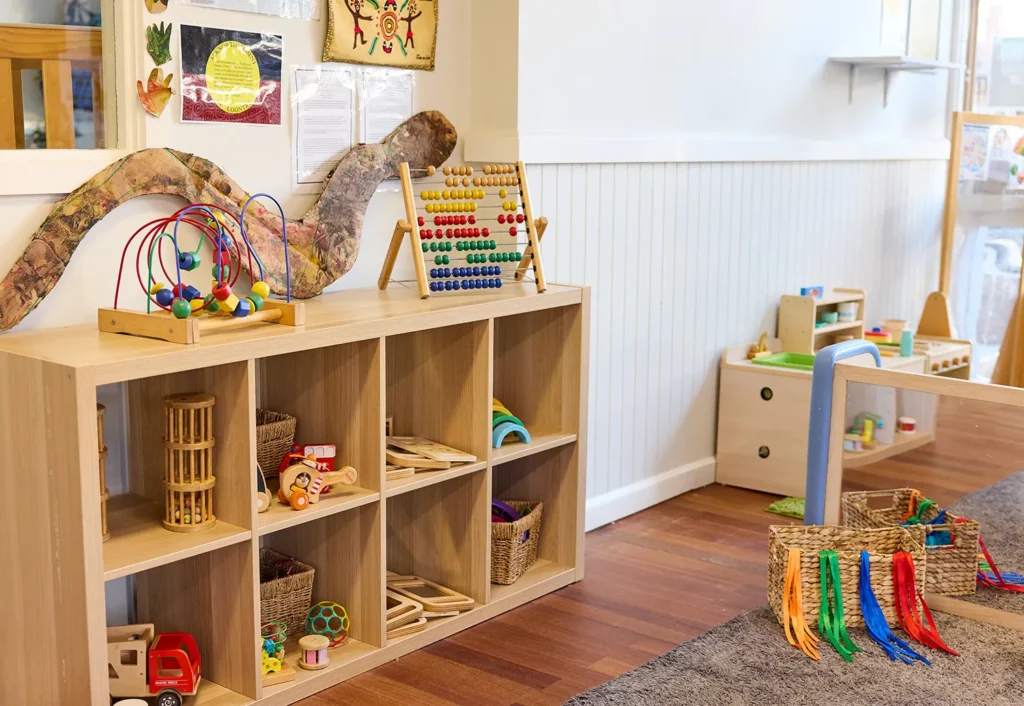 A preschool classroom with wooden shelves containing various educational toys such as an abacus, bead maze, and baskets of blocks. The room has a light, organized, and cheerful atmosphere ideal for early learning.