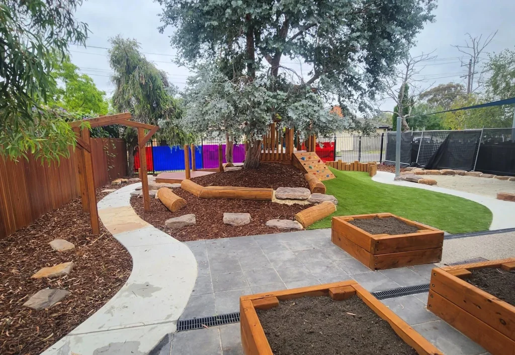 A landscaped outdoor play area at the day care features a climbing structure, wooden planters, mulch, grass, and a paved walkway. Trees and colorful fencing brighten the background, creating an inviting space for children.