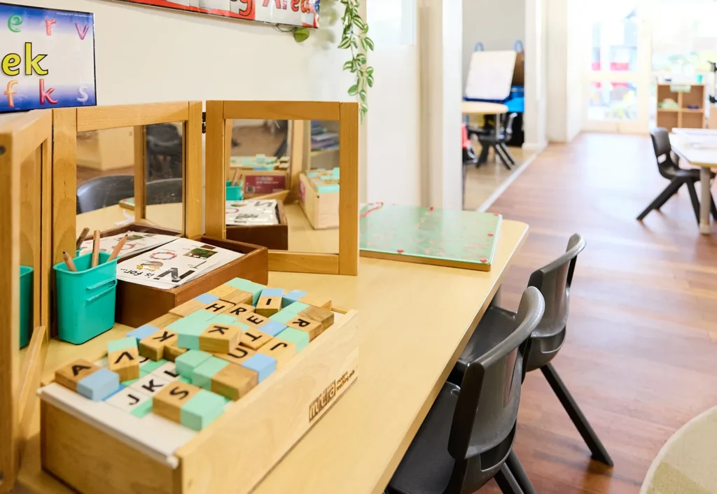 A cheerful preschool classroom with a wooden table holding letter blocks, a wooden mirror stand, and chairs. In the background, more tables and chairs are neatly arranged under large windows that let in abundant natural light.