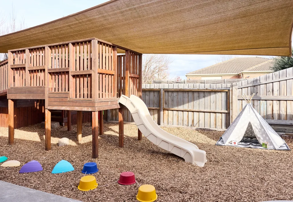 A wooden play structure with a slide stands under a sunshade in a playground, perfect for early learning and childcare. Colorful plastic stepping stones and a white teepee tent are on the mulch ground nearby.