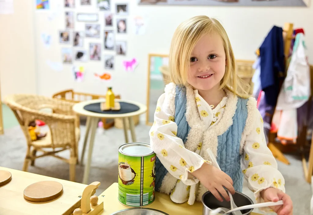 A young child with blonde hair is smiling while holding a spoon next to a can on a small kitchen counter in a preschool playroom. The background shows children's artwork and other play areas.