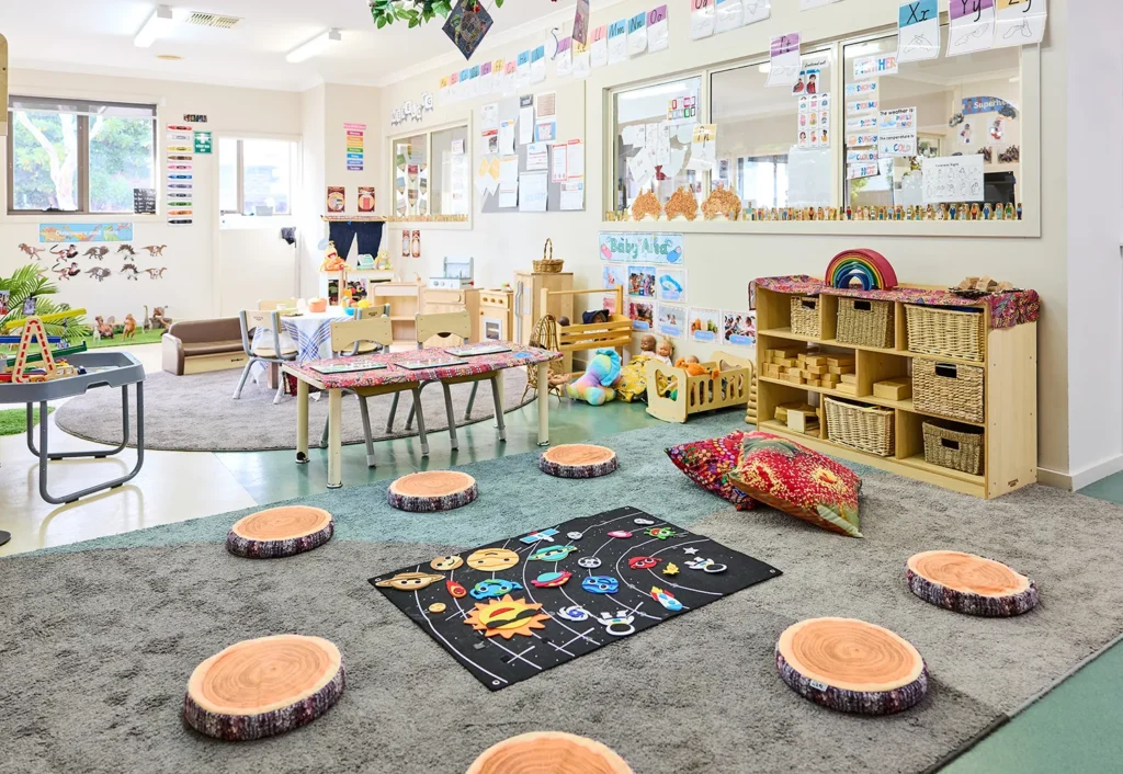 A colorful kindergarten classroom with circular wooden seats, a rug with educational graphics, bookshelves, and various learning materials. Walls are decorated with artwork and educational posters, creating an engaging childcare environment.