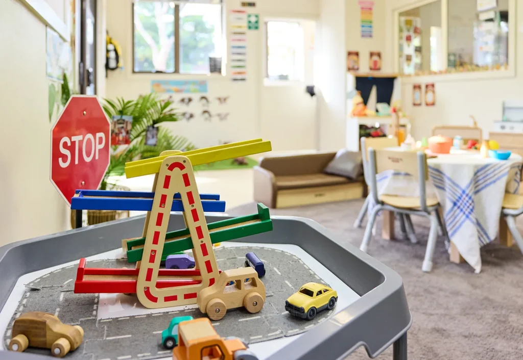 A well-lit room in a kindergarten with a children's play area featuring toy cars, a stop sign, and a ramp on a table. In the background, a table, chairs, couch, and educational posters are visible.