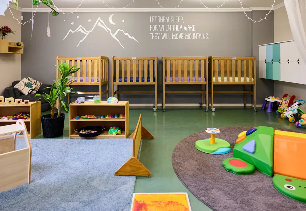 A colorful, well-organized preschool nursery room with cribs, toys, and educational materials. A wall features a painted mountain mural and the quote, "Let them sleep for when they wake they will move mountains." Perfect for childcare and early development.