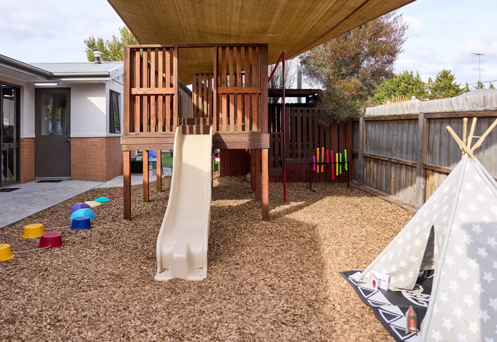 A playground with a wooden climbing structure, slide, and a canopy designed for preschool fun. There's a teepee tent to the right on wood chips and colorful obstacle course pieces to the left, creating an ideal setting for childcare activities.