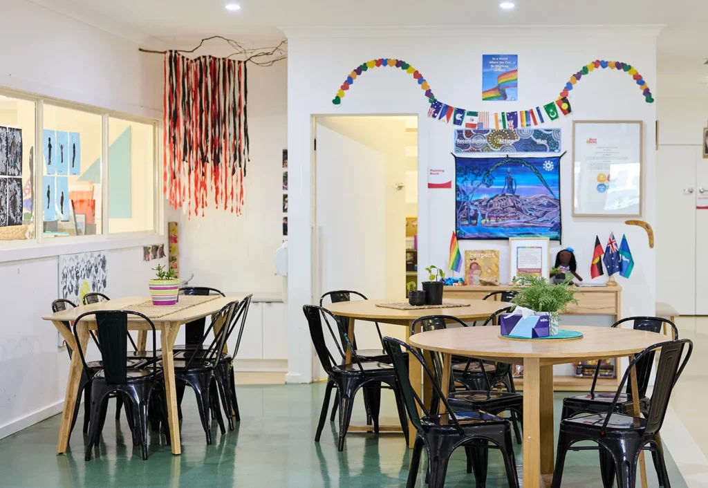 A preschool classroom with round tables and black chairs, decorated with art and crafts on the walls, including a colorful hanging decoration and a "Welcome" banner.