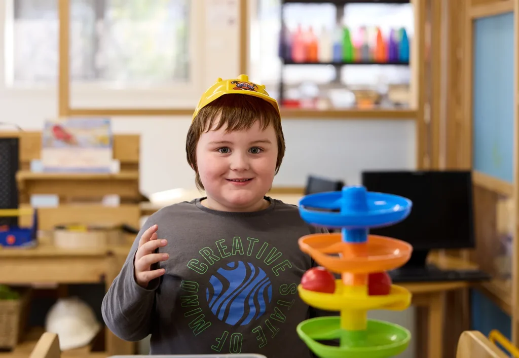 A child wearing a yellow toy hat stands in a day care classroom, smiling and gesturing with hands. In front is a colorful toy with stacked rings. Shelves with books and supplies are in the background.