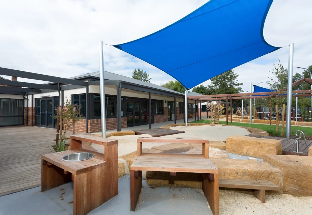 Outdoor play area with wooden benches and tables, sandy ground, and blue shade sails. A modern building and playground equipment are visible in the background.