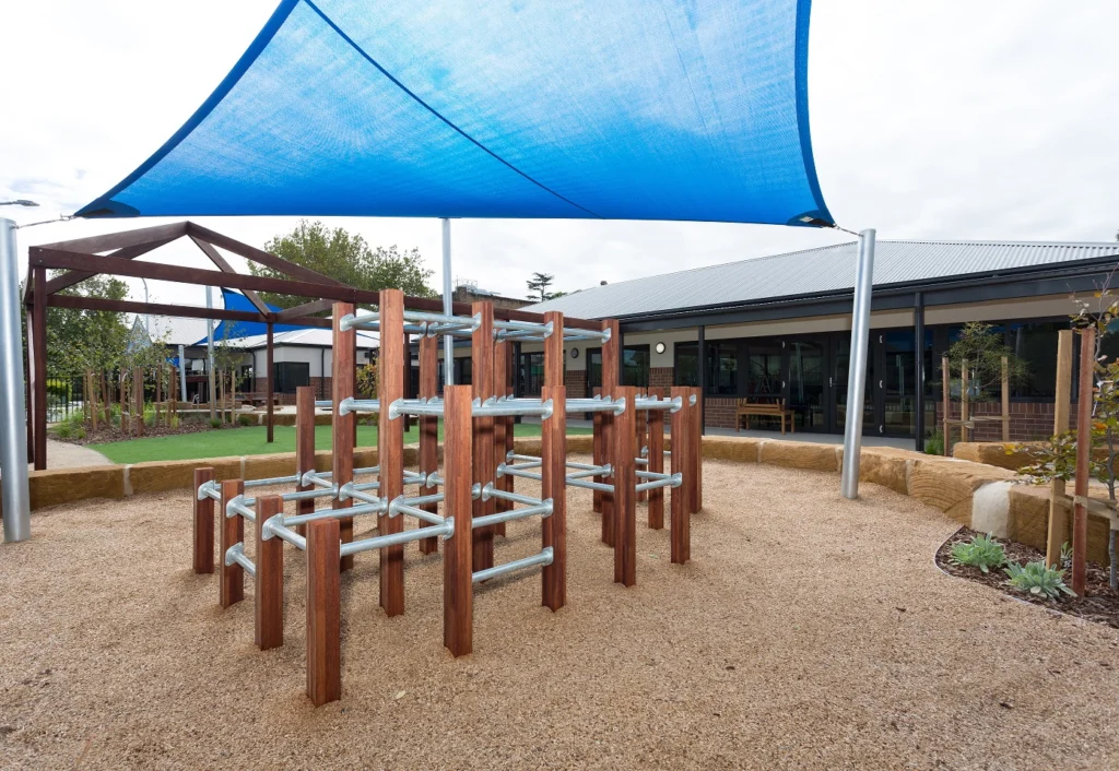 Outdoor play area with a wooden climbing structure and a blue shade sail overhead. There are buildings and greenery in the background.