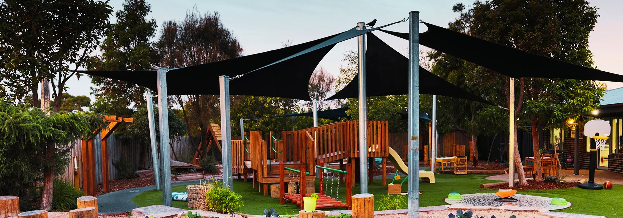 An outdoor playground at Busy Bees Narre Warren childcare featuring wooden play structures, climbing equipment, and black sunshades.