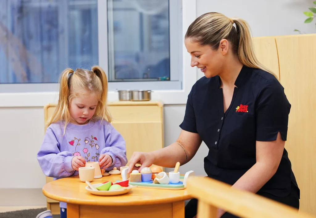 A young girl and a woman sit at a small table, playing with toy kitchen items. The girl is wearing a purple shirt, and the woman is dressed in black with her hair tied back.