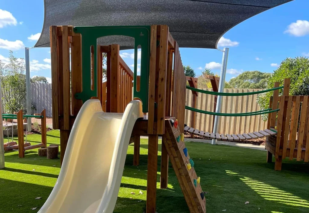 A children's playground set with a slide, climbing rocks, and shaded areas on an artificial grass surface. A wooden fence and some trees are visible in the background.