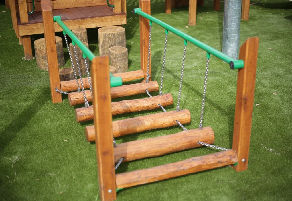 A wooden log bridge with chain supports is set up on a playground with artificial turf ground.