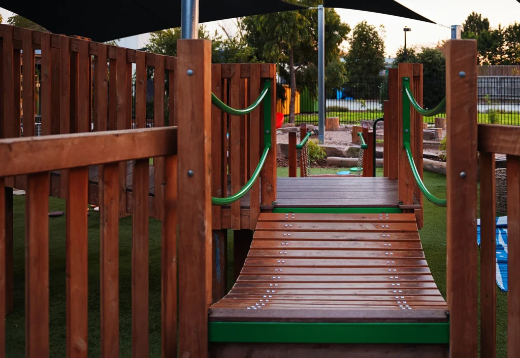 Wooden playground bridge with green handrails, connecting platforms. Shade structure overhead, trees, and playground equipment visible in the background.