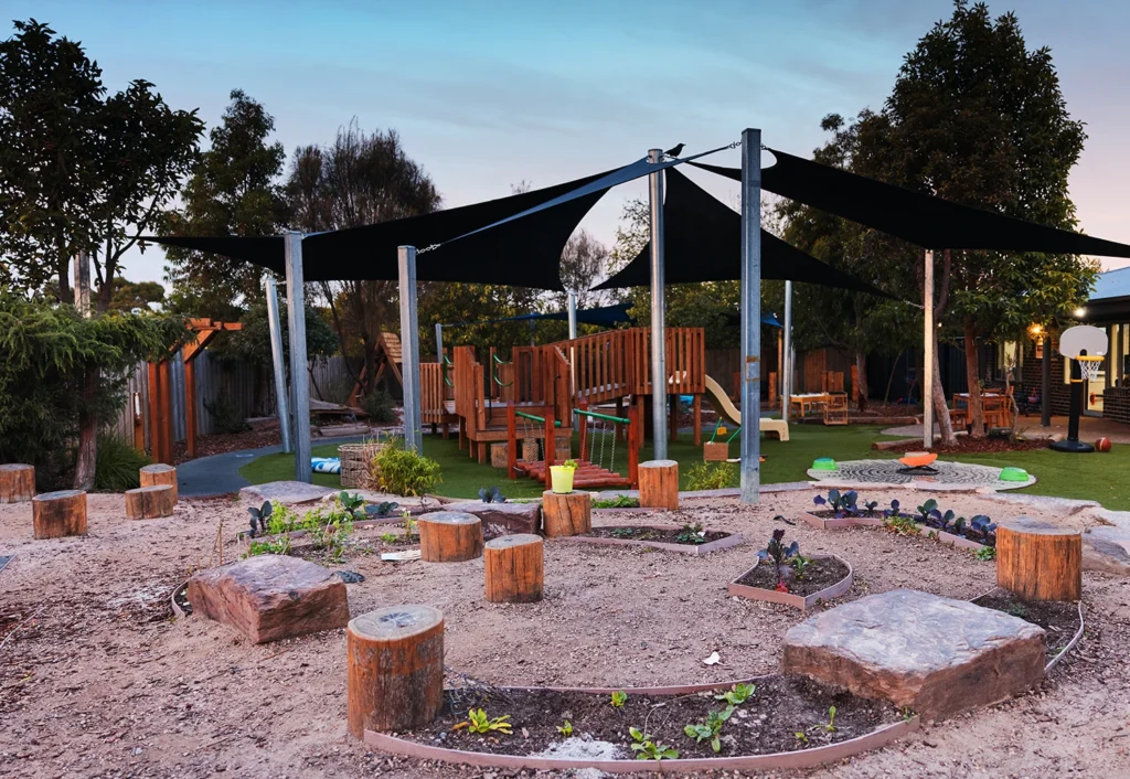 Children's playground with wooden climbing structures, slides, sand area with tree stumps, a garden, and shade sails. Trees and fencing surround the play area.