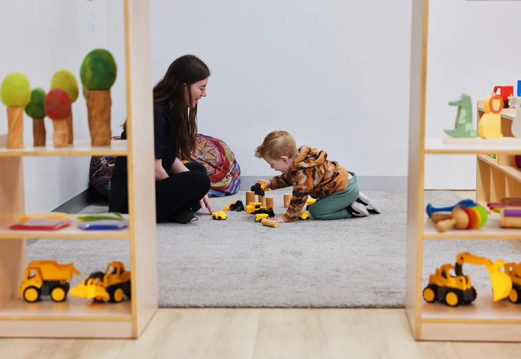 A woman and child are sitting on a carpet playing with toy construction vehicles. Shelves with toys and items frame the scene.