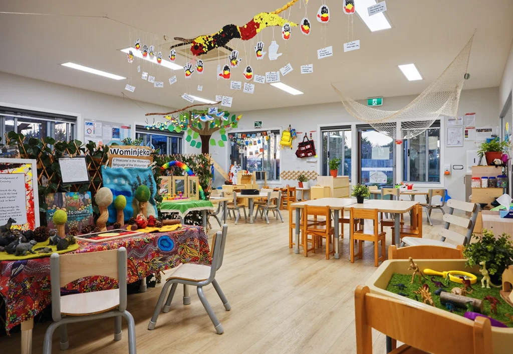 A colorful classroom with various educational displays, child-sized furniture, and a vibrant ceiling with hanging decorations. Shelves and tables are filled with learning materials and toys.