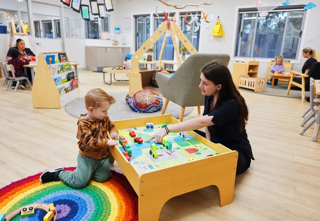 A young child and an adult play with toy cars on a table in a brightly lit classroom. Other children and adults are engaged in various activities in the background.