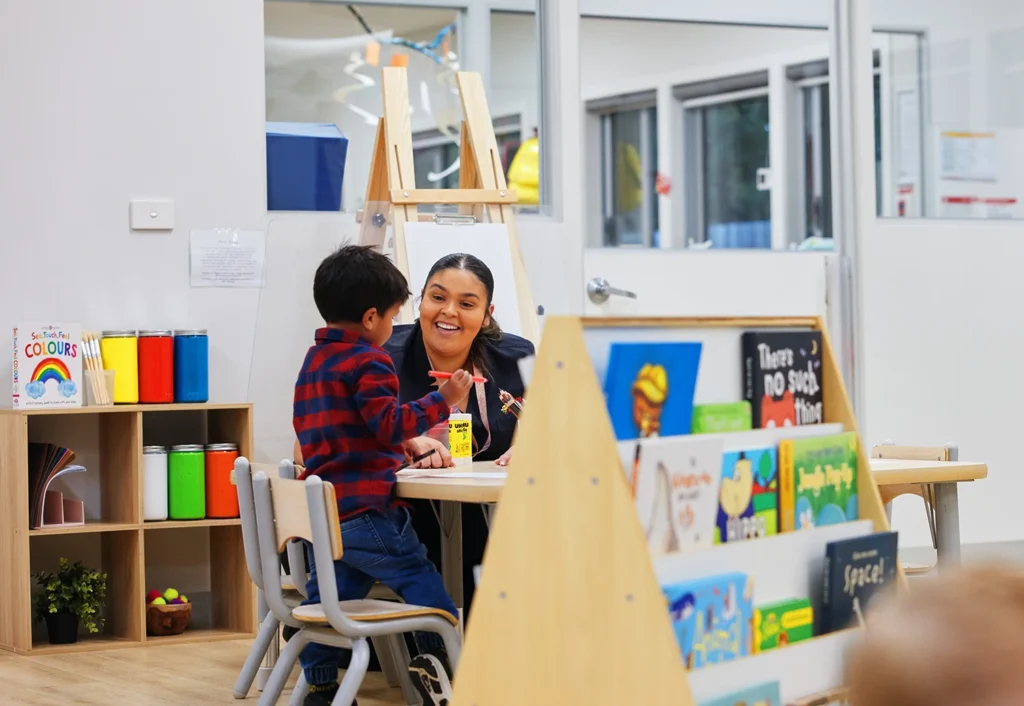 A teacher and a young student interact at a small table in a classroom. Nearby, colorful books and art supplies are visible on shelves.