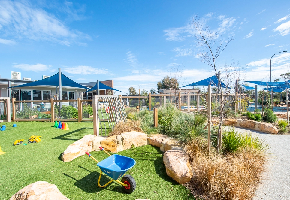 A playground with artificial grass, blue shade structures, scattered rocks, and various play equipment including a wheelbarrow is shown on a sunny day. A building is visible in the background.