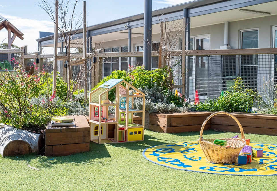 Outdoor play area featuring a wooden dollhouse, a wicker basket with toys, and a circular play mat on a grassy surface. The area is surrounded by plants and a building in the background.