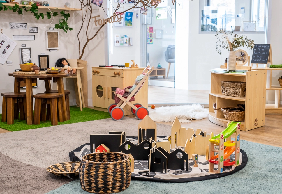 A bright and organized preschool classroom features wooden furniture, educational toys, a play kitchen, a wooden doll stroller, and a play mat with toy buildings and structures.