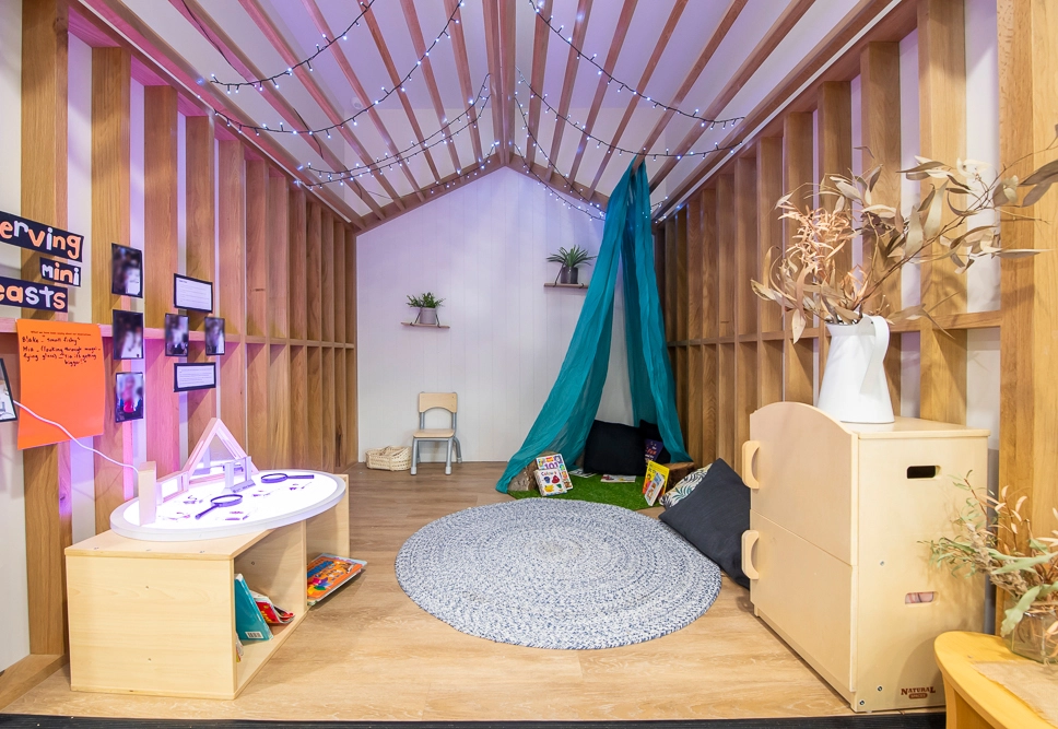 A cozy reading nook with a rug, cushions, a small table, and a tepee. Wooden walls are adorned with fairy lights, plants, and children's books.