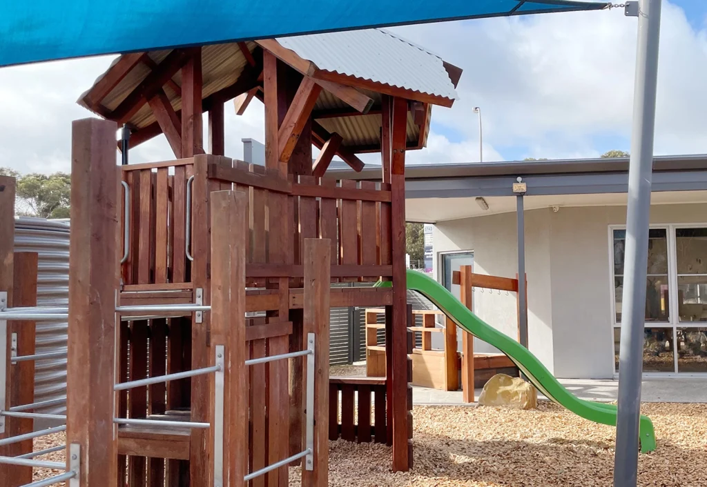 A playground at Noarlunga Downs childcare featuring a wooden climbing structure with a slide, under a shade canopy.