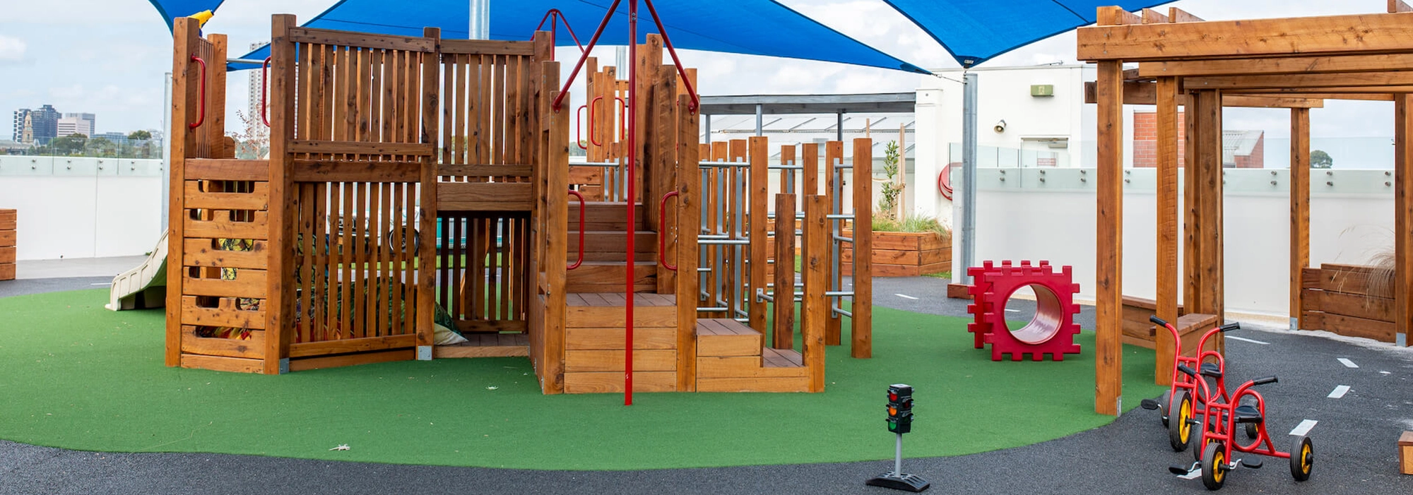 An outdoor playground features wooden structures with slides, climbing equipment, and a small red tricycle. Blue shade sails are overhead, and the ground is a mix of green turf and blacktop.