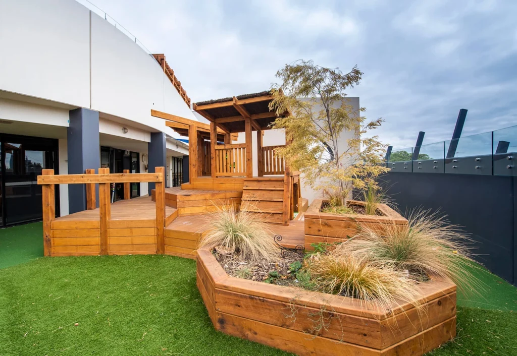 A rooftop garden with wooden planters and walkways, featuring a small pergola and various plants. The area is surrounded by glass barriers and overlooks an urban setting on a cloudy day.