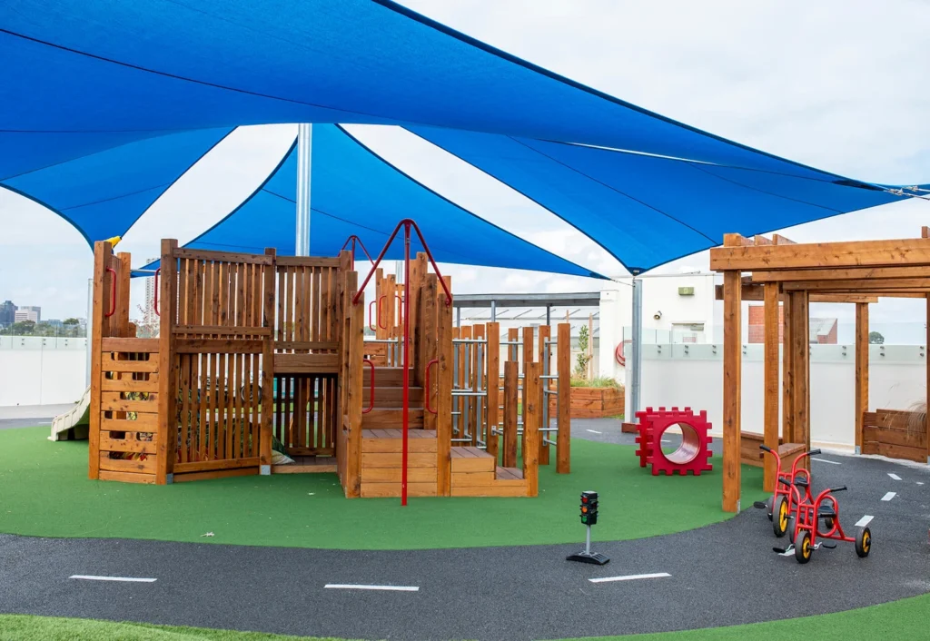 A playground with wooden structures, including slides and climbing equipment, is covered by blue shade sails. The ground has green artificial turf and a tricycle path. Red tricycles are parked nearby.