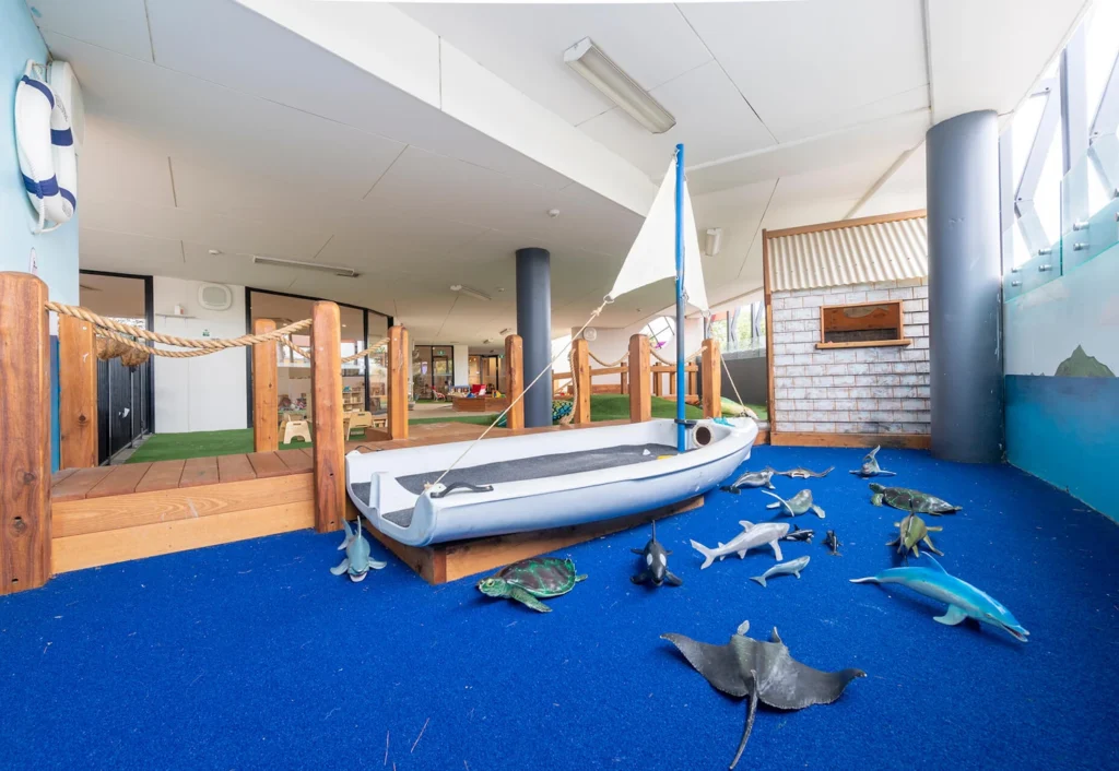 Indoor play area with a boat, blue carpet, and various toy sea animals. The space includes a wooden fence, a bridge, and large pillars.