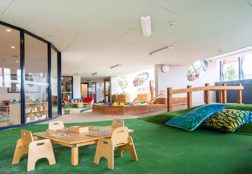 A brightly lit and spacious children's play area with toys, small tables with chairs, and a wooden play structure on artificial turf. A large glass wall reveals an adjacent indoor space.