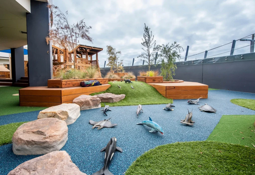 Outdoor play area with artificial turf, large rocks, and model animals, including dolphins and sharks, on a blue rubber surface. Wooden seating and planters are present in the background.