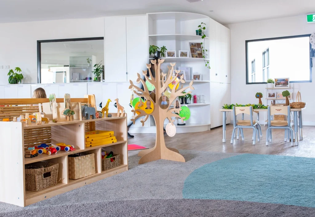 A bright and organized classroom features a wooden toy tree, shelves with various toys, small tables, and chairs. Large windows let in natural light, creating an inviting learning environment.