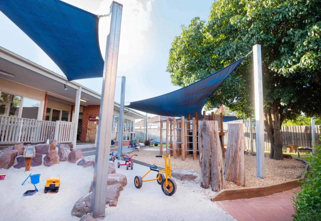 Outdoor playground with climbing structures, a shaded play area, toy vehicles, and sandy ground. Trees and building in the background.