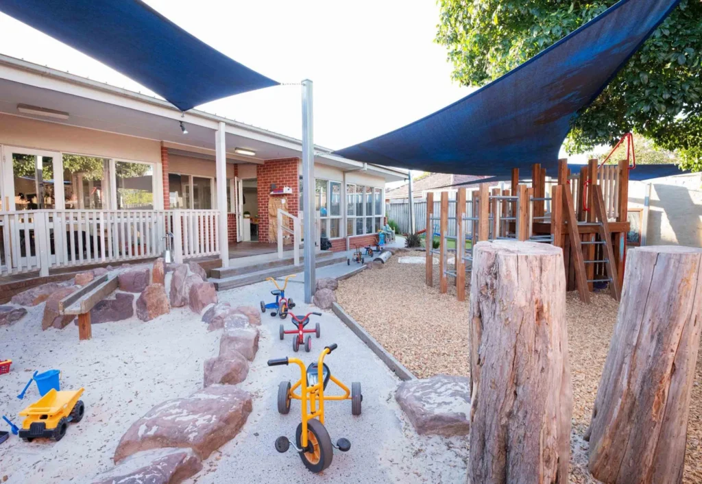 An outdoor play area with tricycles, toy trucks, a sandpit, climbing structures, and shaded blue canopies at a childcare center. Wooden logs and benches are spaced throughout.