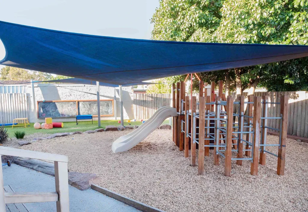 A playground with a slide, climbing structure, and shaded area on wood chips. There is a bench and fence in the background and a large blue shade above.