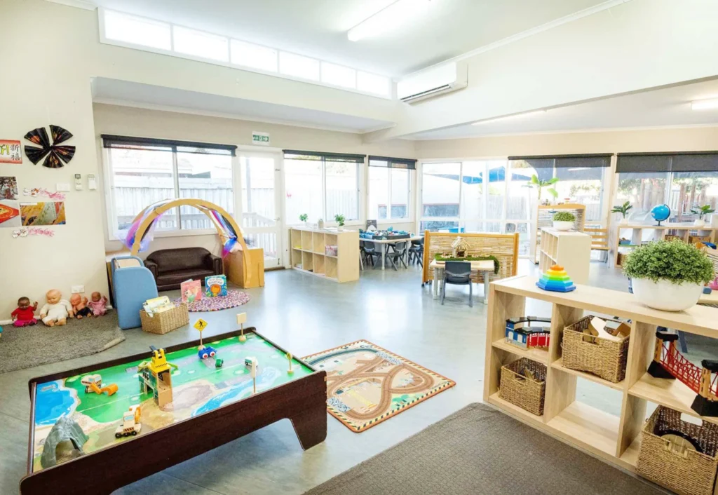 A well-lit, spacious classroom with various educational toys and activities. Shelving units hold baskets and materials, while tables and chairs are arranged for group activities. Large windows offer ample natural light.