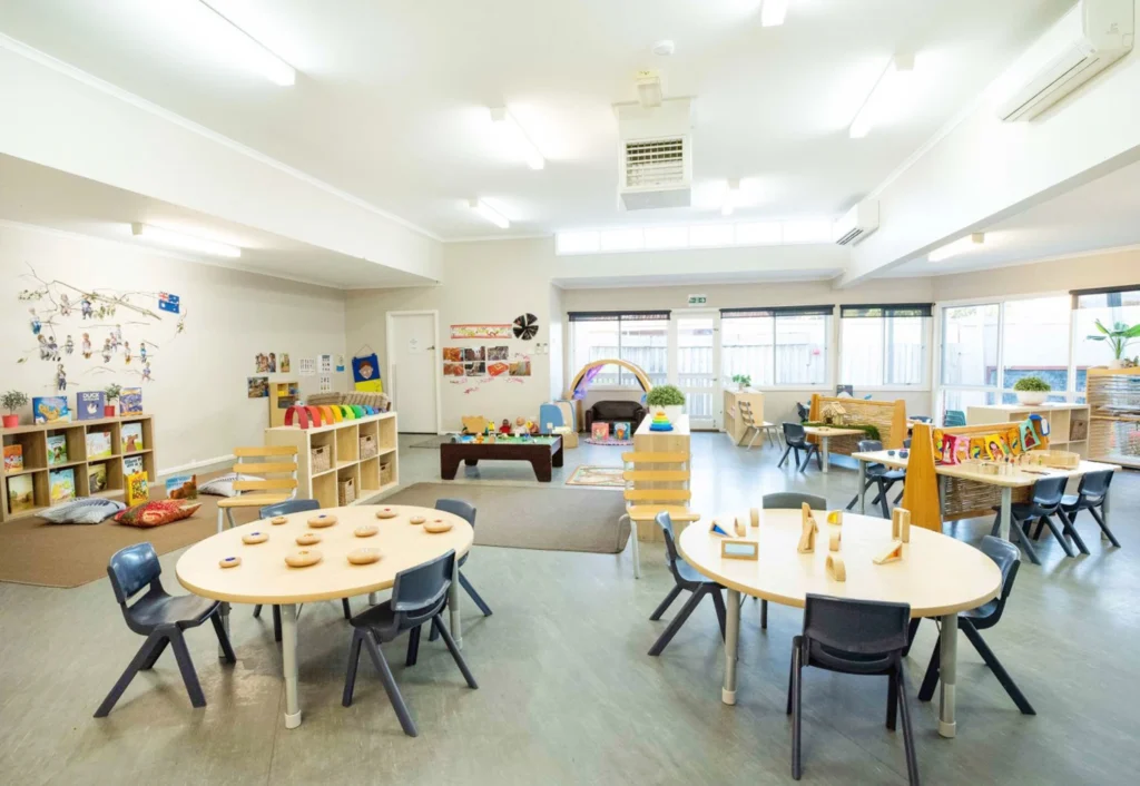 A bright, spacious classroom with several activity tables and chairs. Various educational materials and toys are neatly arranged on shelves and tables throughout the room. Large windows provide natural light.