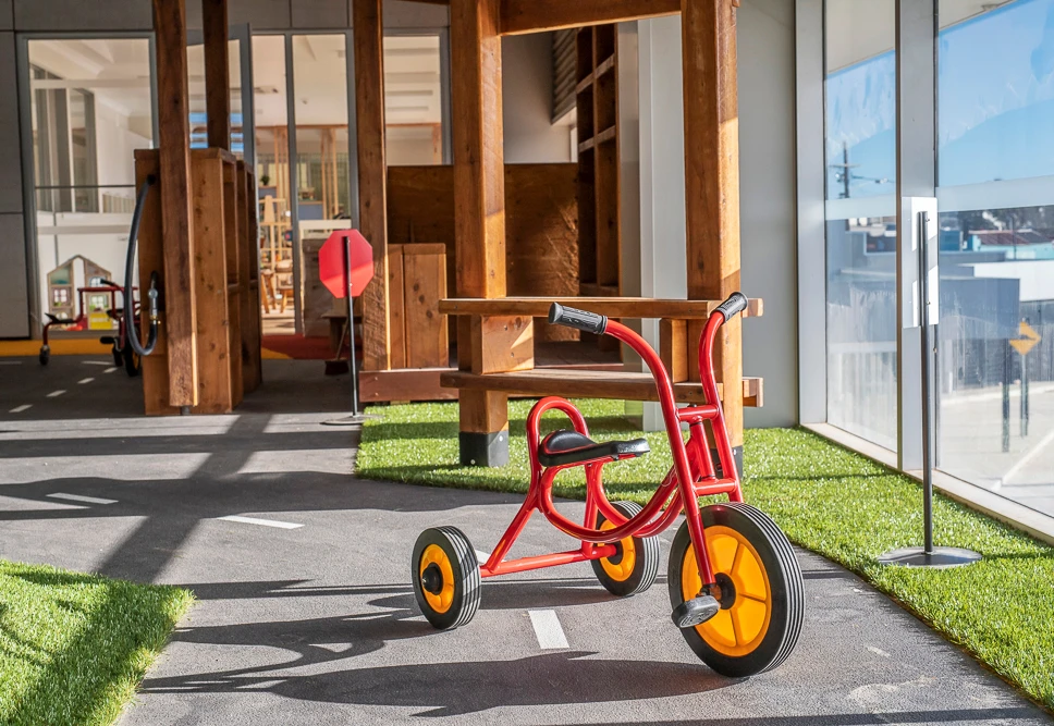 A red tricycle with yellow wheels is parked on a mini roadway inside a play area, which has artificial grass and wooden structures.