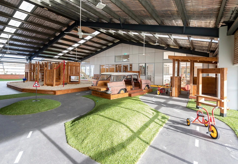 Indoor playground with various play structures, including a wooden playhouse, a sandbox with a climbing frame, a vintage car model, and a tricycle on a mini road track with markings.