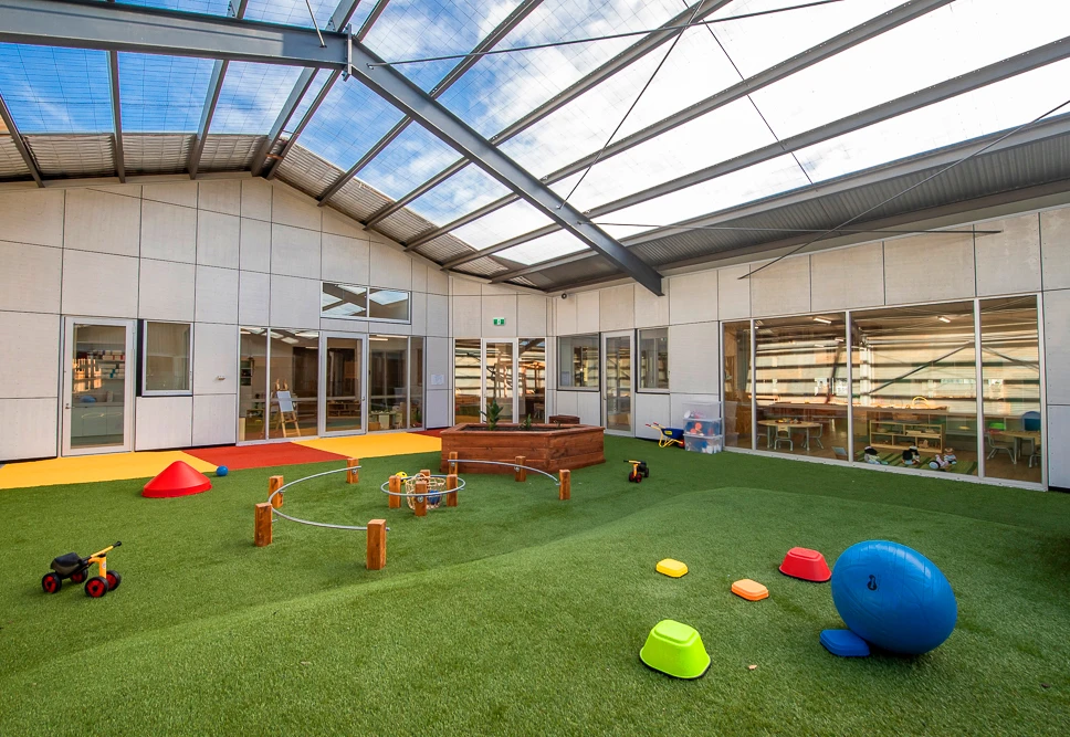 A bright, spacious indoor play area with artificial turf, various colorful playground equipment, a sandbox, and large windows under a high, transparent ceiling.