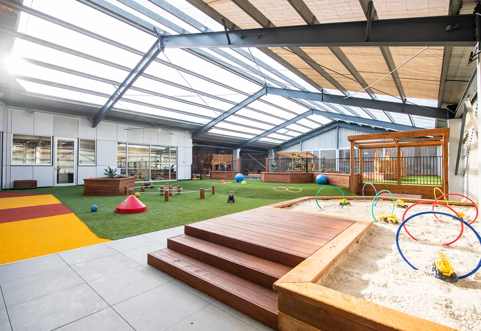 Indoor playground with artificial turf, sandpit, and various play equipment, including a wooden climbing structure, seating areas, and colorful hoops. The area is under a large translucent roof.