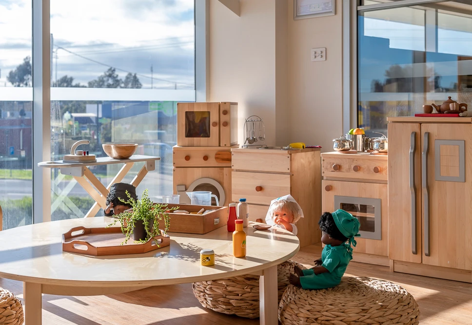 A well-lit playroom with toy kitchen set, dolls, and various play items on a round table and shelves. Large windows in the background let in natural light.