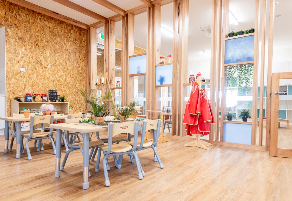 A brightly lit kindergarten classroom at Shepparton childcare centre with wooden walls and floors, tables and chairs, greenery, and children's red art aprons on a stand.