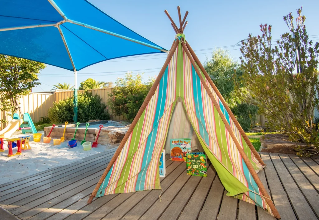 A striped teepee stands on a wooden deck beside a sandpit with sand toys under a large blue shade umbrella in a backyard, creating an ideal space for early learning. Children's books and cushions are inside the teepee, perfect for kindergarten storytelling sessions.