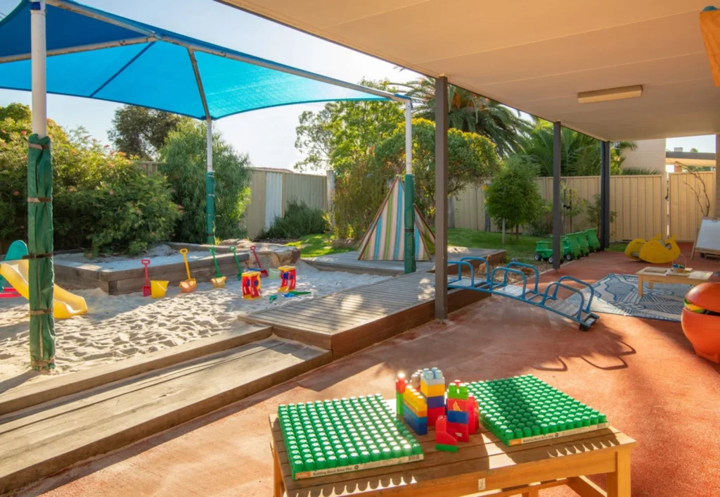 A covered outdoor play area with a sandbox, colorful toys, a slide, and seating. Perfect for preschool activities or an early learning environment, a blue shade structure provides protection from the sun. The area is surrounded by greenery and a secure fence.