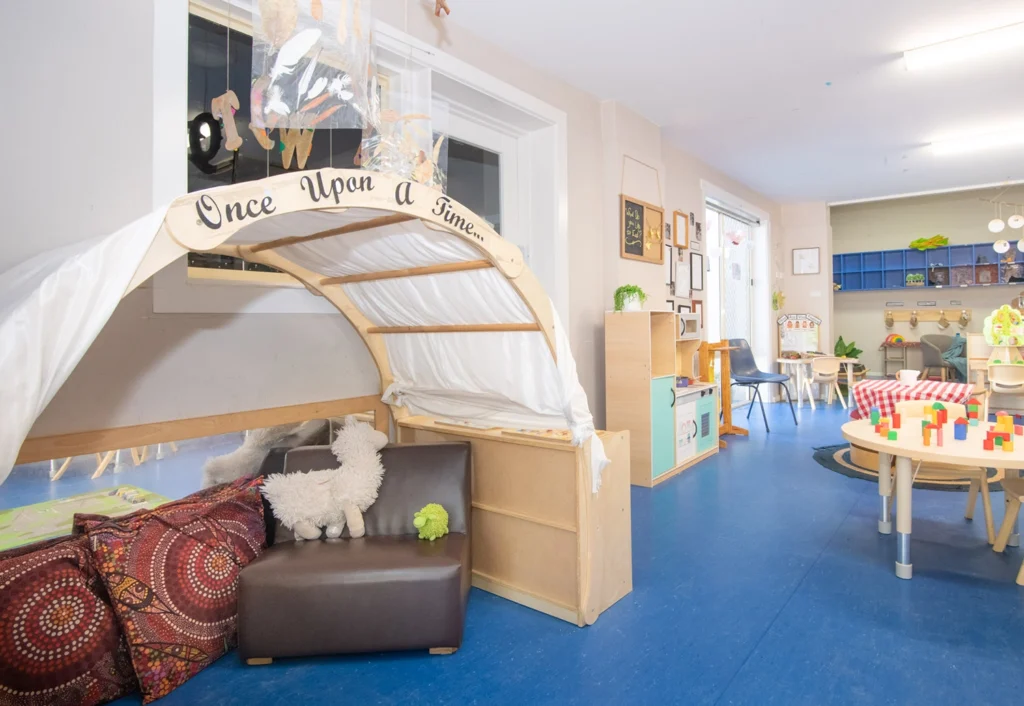 A colorful kindergarten classroom features a "Once Upon a Time" reading nook, toys, tables with chairs, and blue flooring.