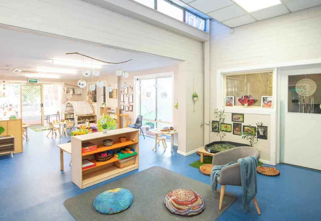 A brightly lit, spacious kindergarten classroom with blue flooring, low shelves stocked with educational materials, colorful cushions on the floor, and large windows letting in natural light.